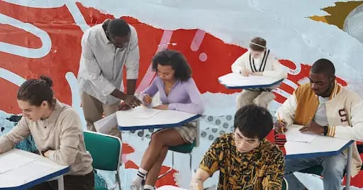 Five teens sit at their classroom desks working on homework while a teacher helps one of them. The scene is on a colorful newsprint background.